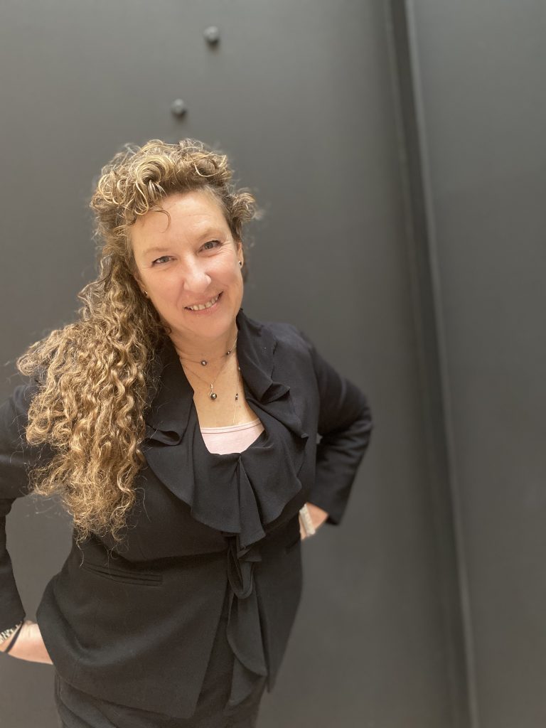 A woman in a black outfit smiles at the camera. Her hands are on her hips.