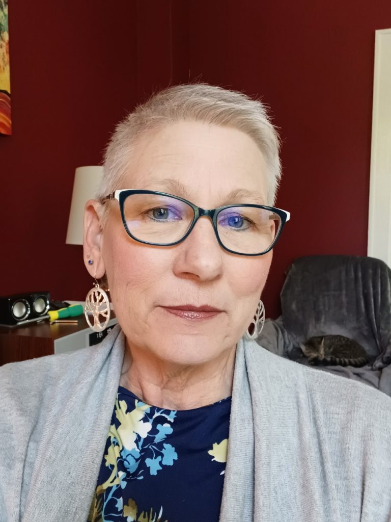 Cynthia is sitting in a living room looking at the camera through black glasses. Her hair is white and worn short and she is wearing tree of life earrings. 